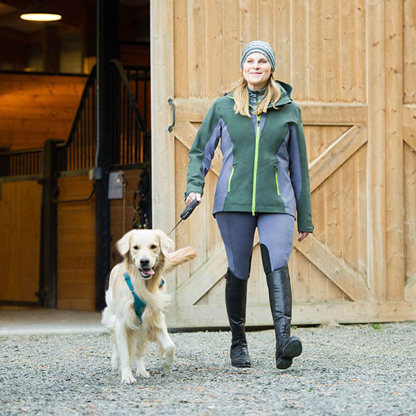 Woman in equestrian apparel walking a dog on a leash by a barn